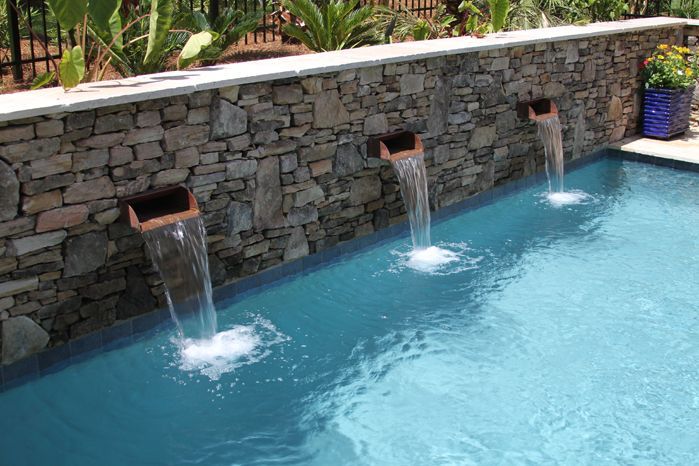 Three waterfalls are coming out of a stone wall into a swimming pool