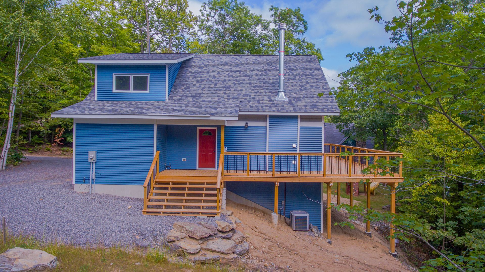 A blue house with a red door and a large deck surrounded by trees.