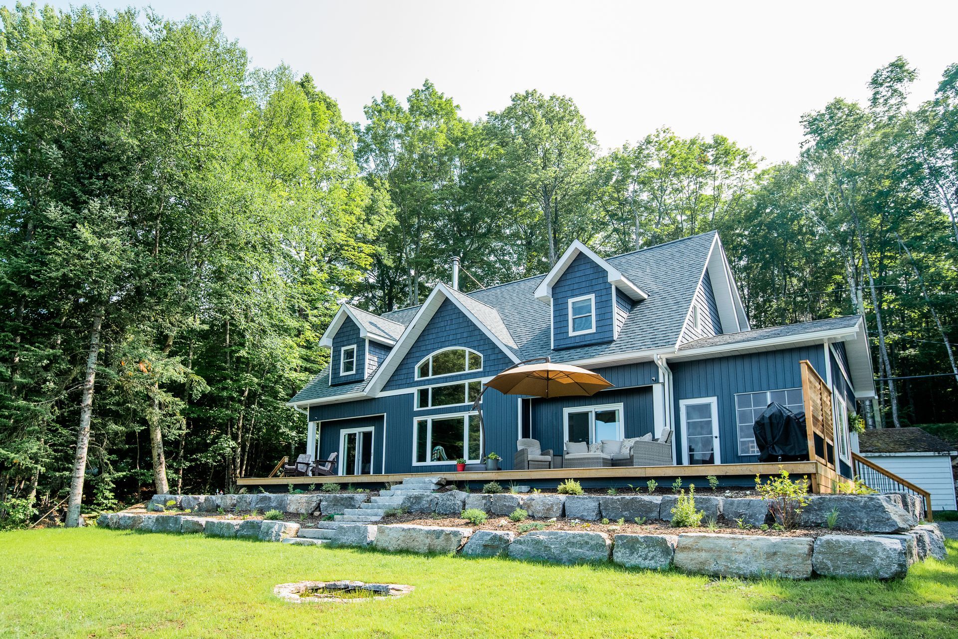 A large blue house with a fire pit in front of it is surrounded by trees.