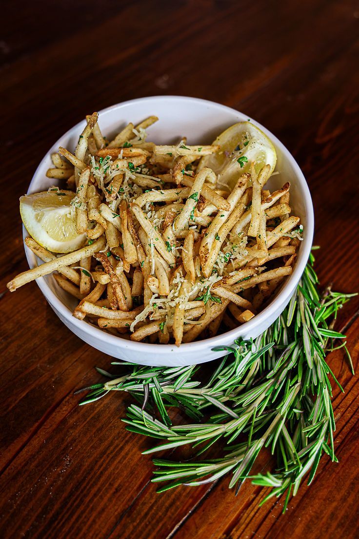 A bowl of french fries with a lemon wedge and rosemary on a wooden table.