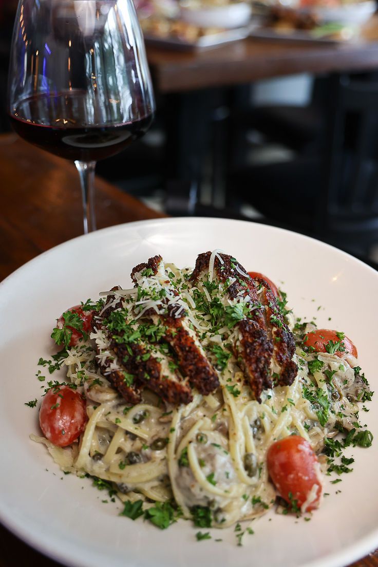 A plate of pasta with chicken and tomatoes next to a glass of wine.
