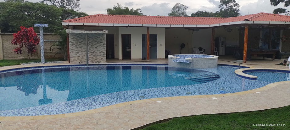 Piscina y jacuzzi junto a una casa con techo rojo. Jardines con vegetación. Cielo nublado.