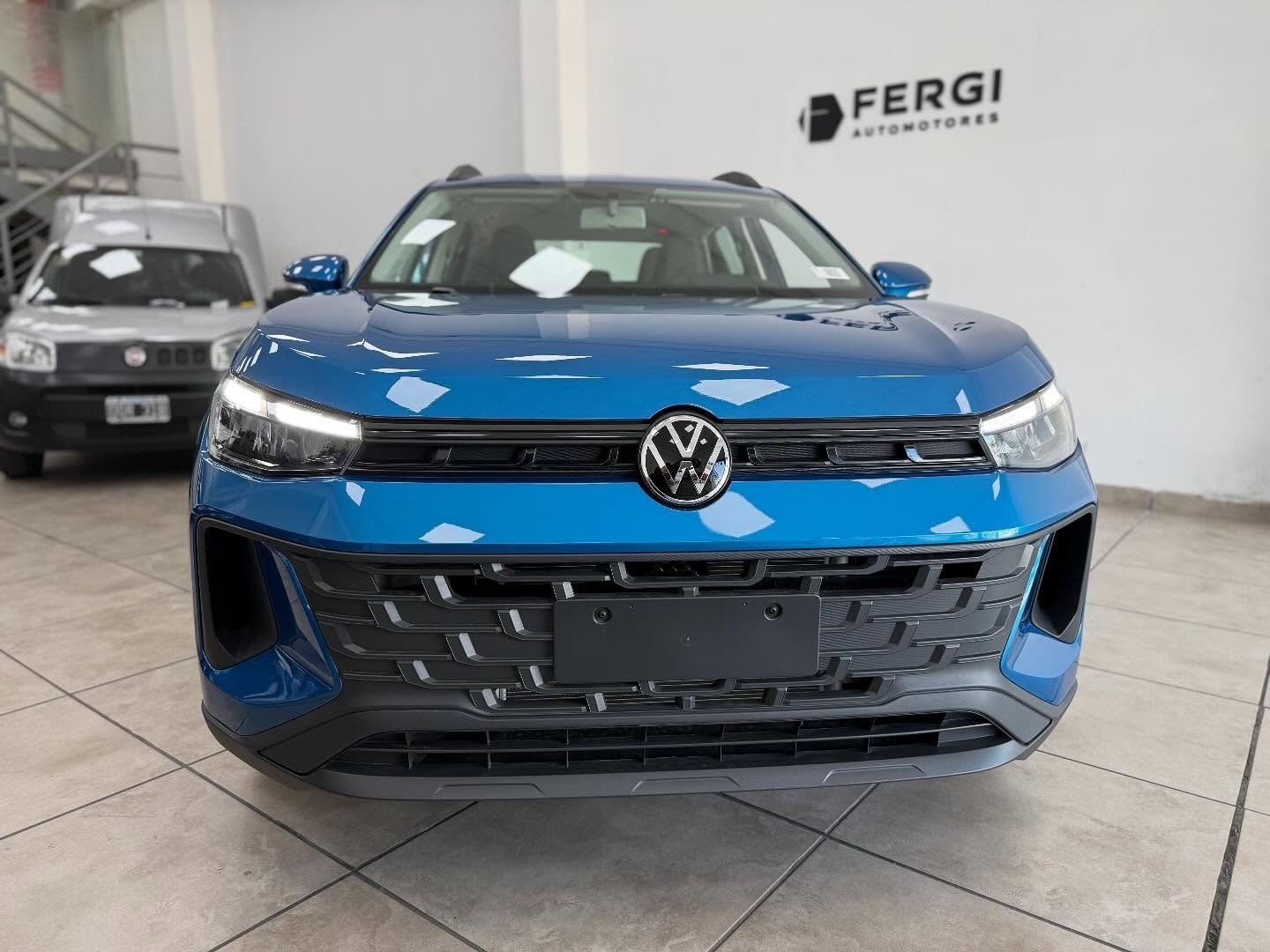 SUV Volkswagen Taos azul estacionado dentro de un concesionario, con un auto plateado al fondo.