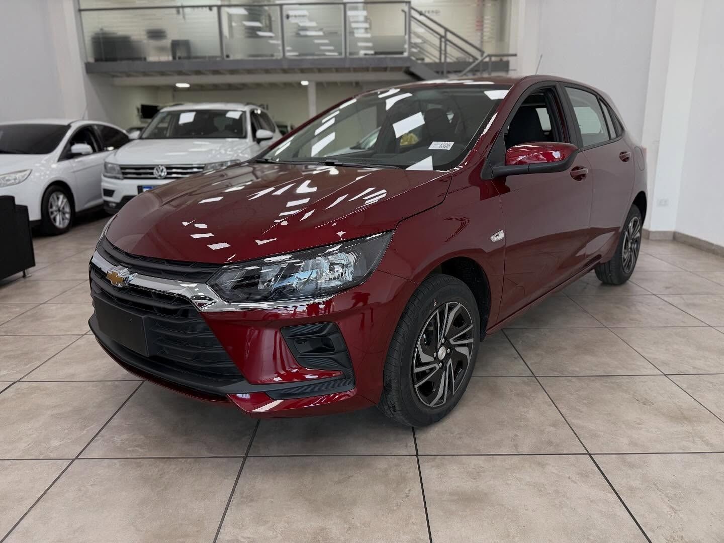 Chevrolet Onix hatchback de color rojo en una sala de exposición, con un Volkswagen blanco y otro coche al fondo.