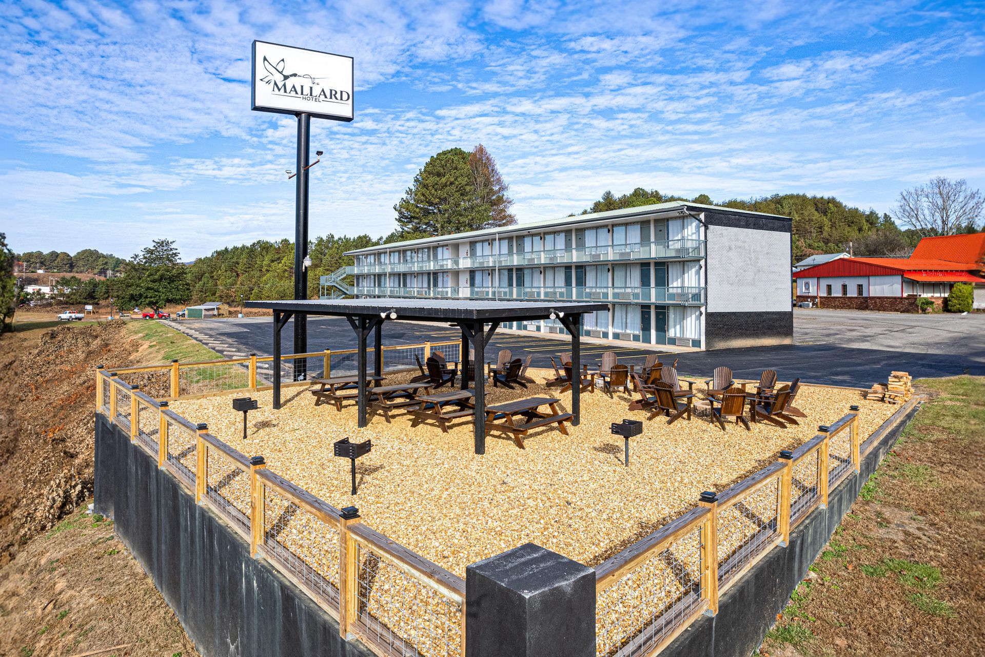 A motel with a sign, picnic tables, and chairs on a gravel platform; sunny day.