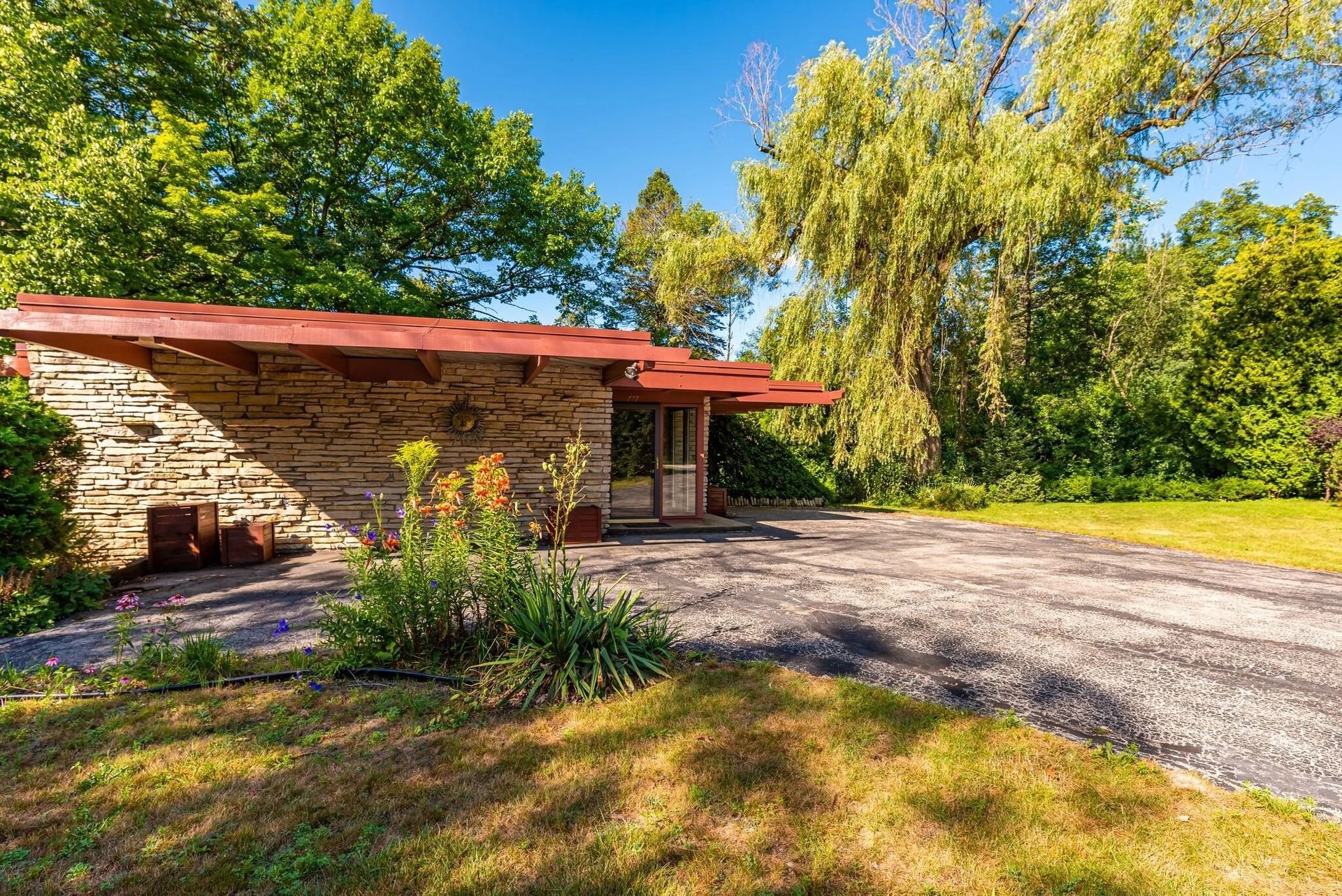Stone house with red roof beside gravel driveway, surrounded by green trees and sunny yard