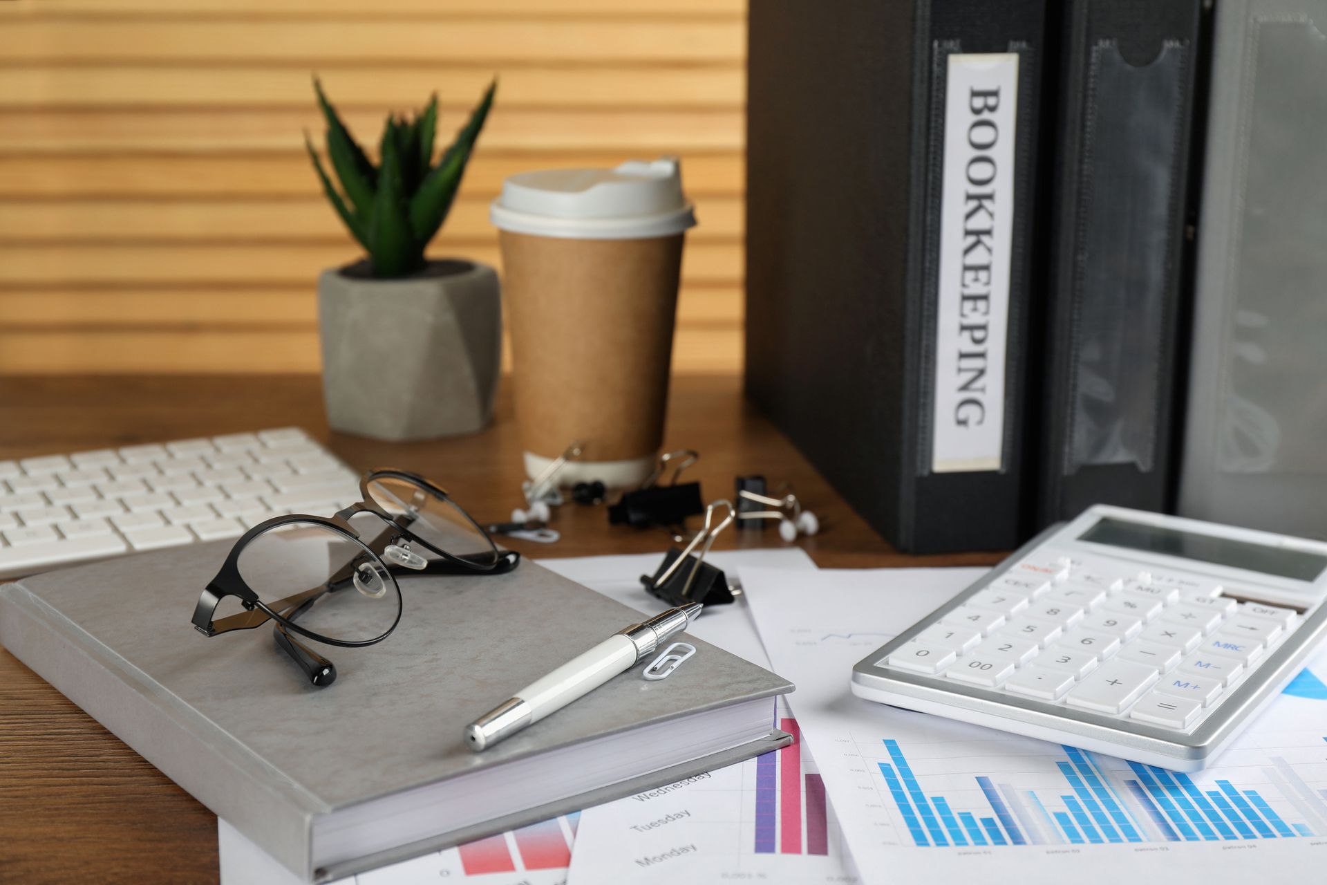 Bookkeeping desk with a notepad, calculator, glasses, coffee cup, and binder.