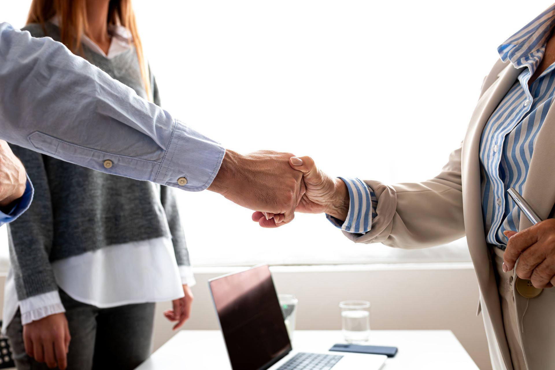 Two people fist bumping in a bright office; third person in background, laptop, water glass on desk.