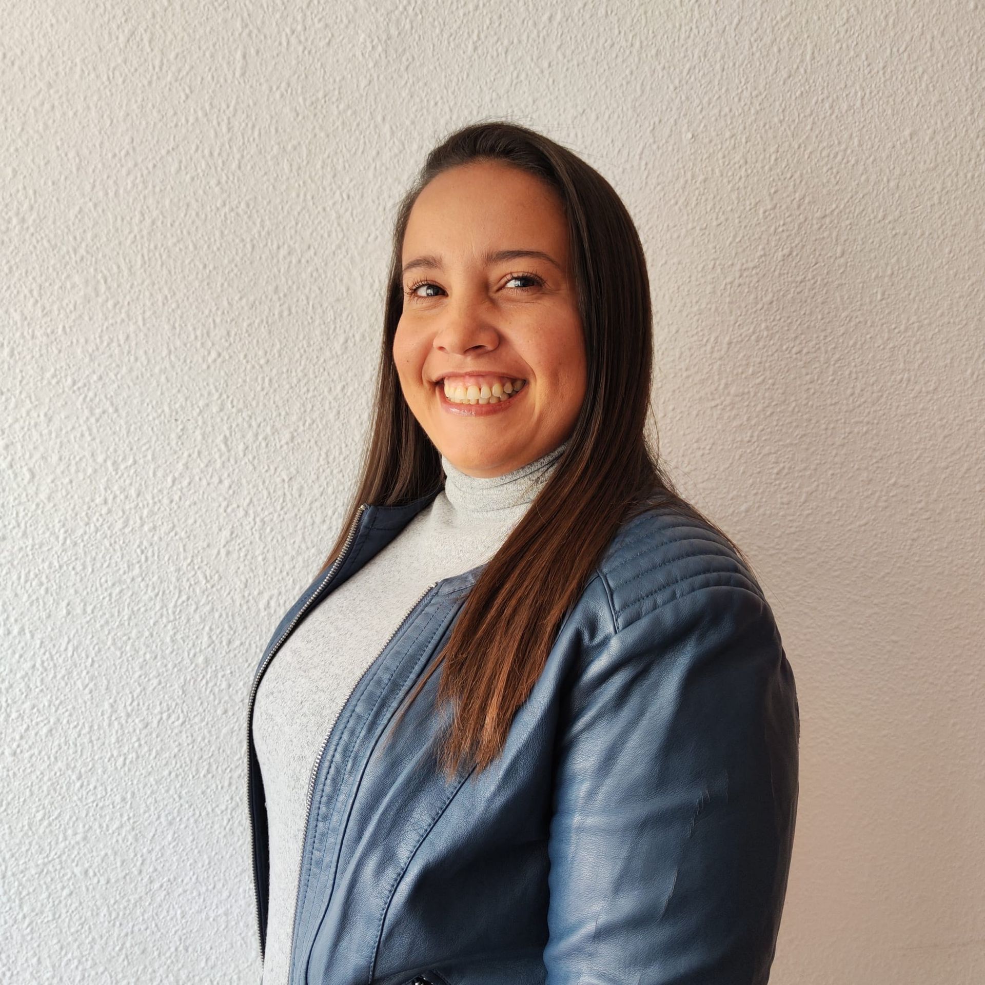 Woman smiling, wearing a gray turtleneck and blue jacket, standing against a white wall.