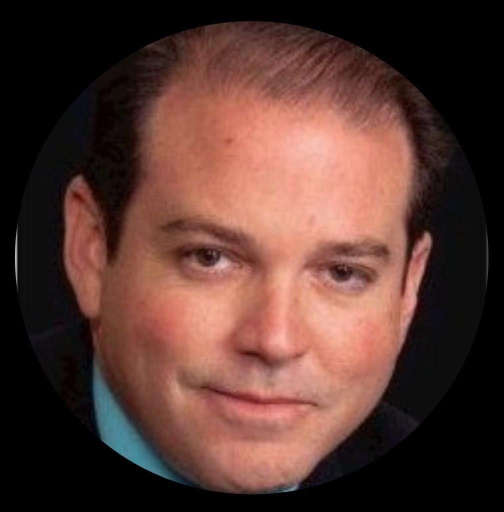 Man with short, dark hair, wearing a suit jacket over a teal shirt, smiles at the camera.
