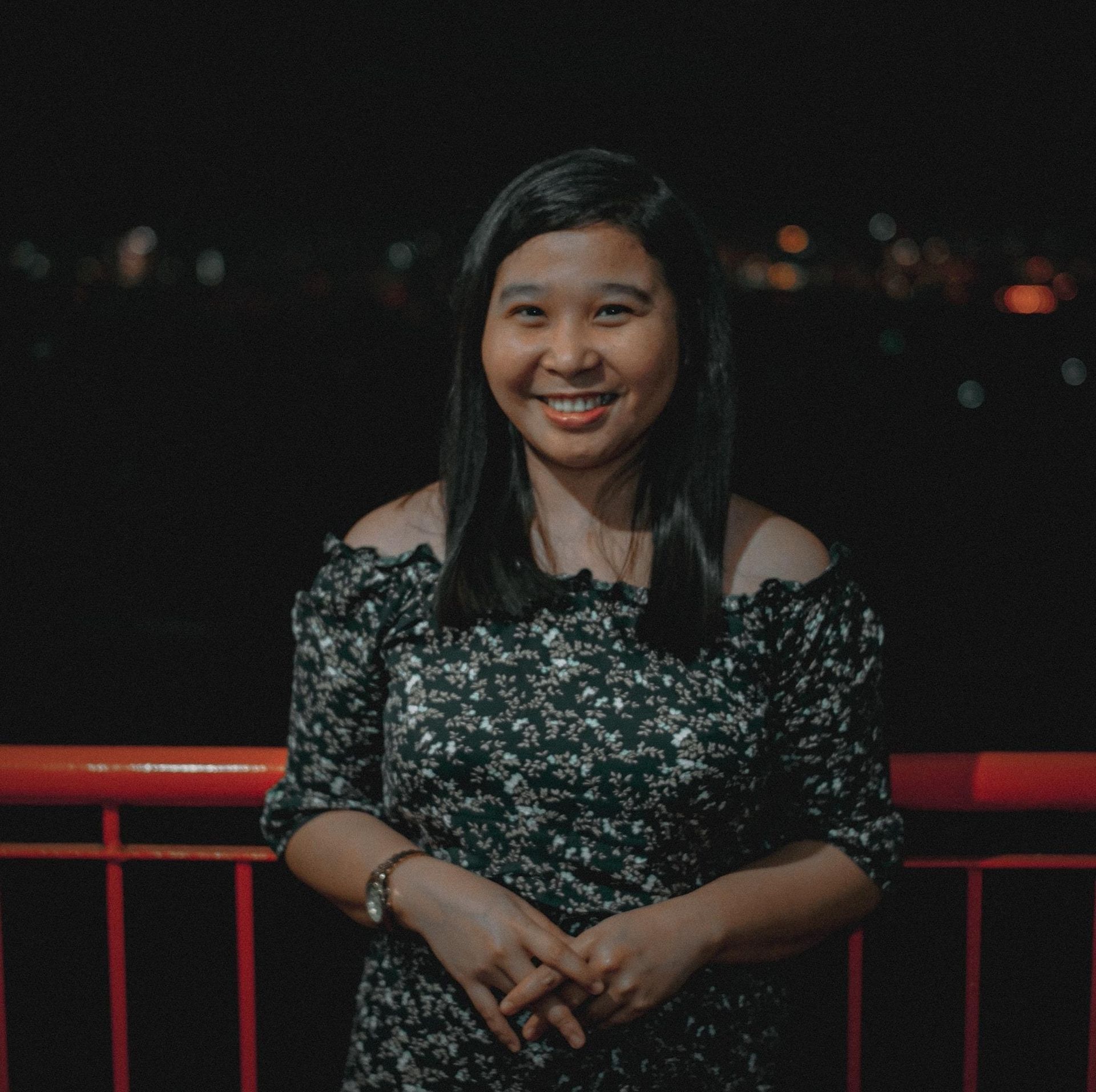 Woman smiling outdoors at night, wearing a floral off-the-shoulder dress. City lights in background.