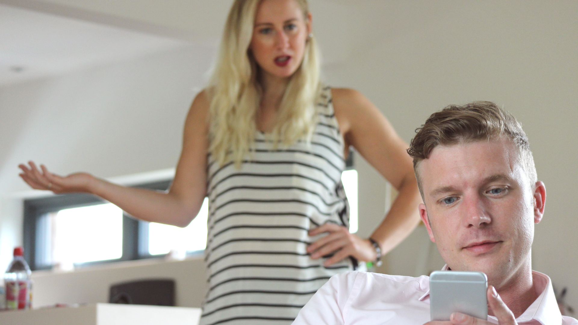 A man is sitting on a couch looking at his cell phone while a woman stands behind him.