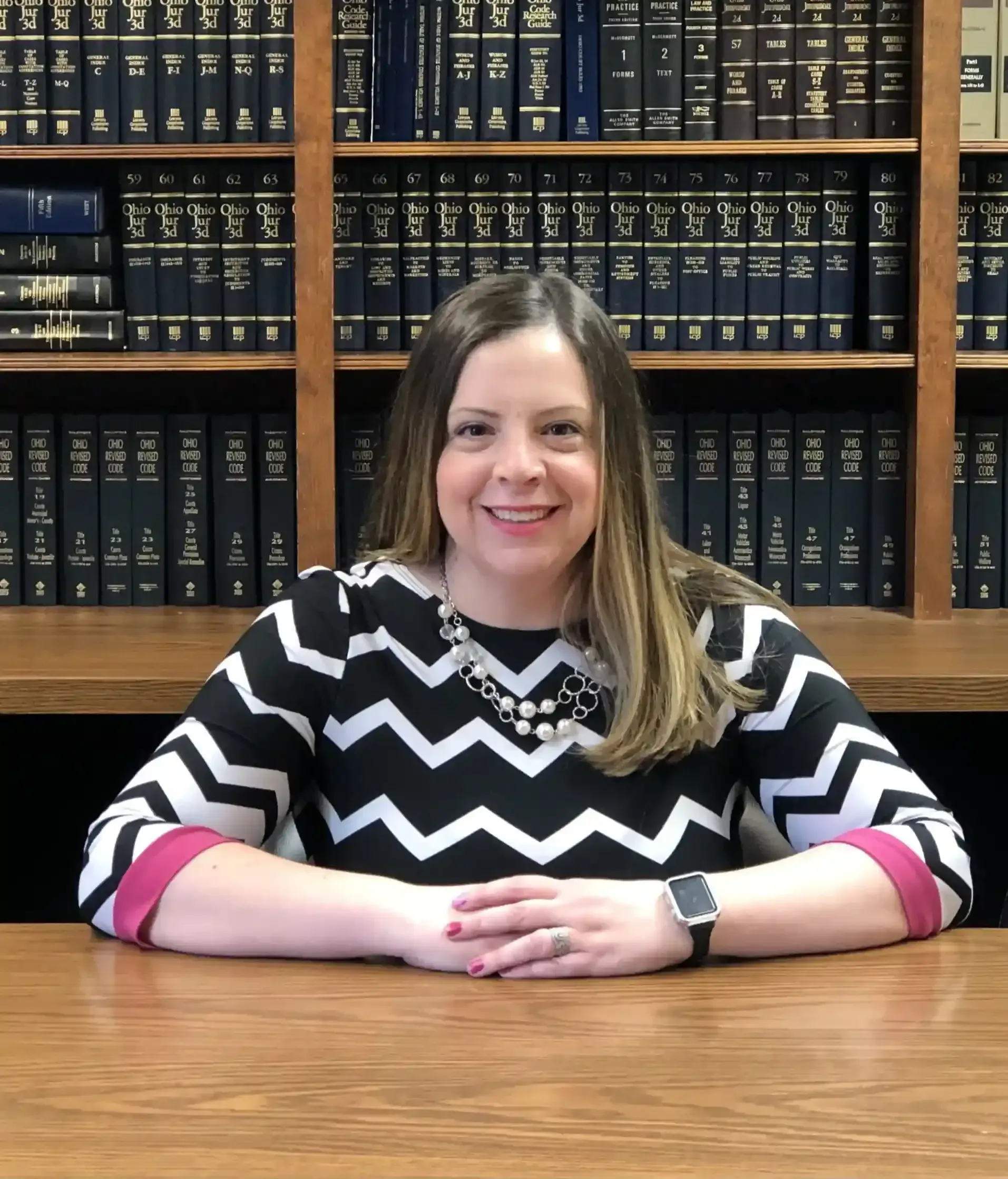 Woman in black and white chevron sweater smiles, sitting at a wooden desk with a library bookshelf background.