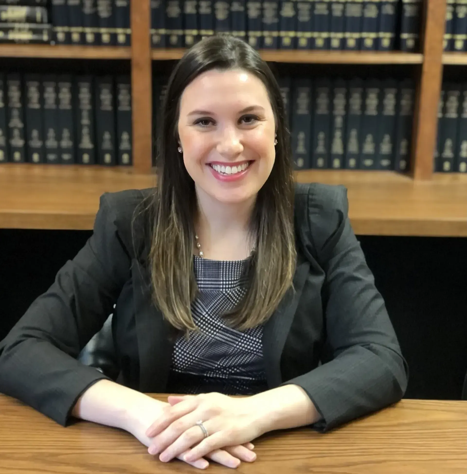 Woman in a blazer smiles at the camera, sitting at a desk with a library backdrop.