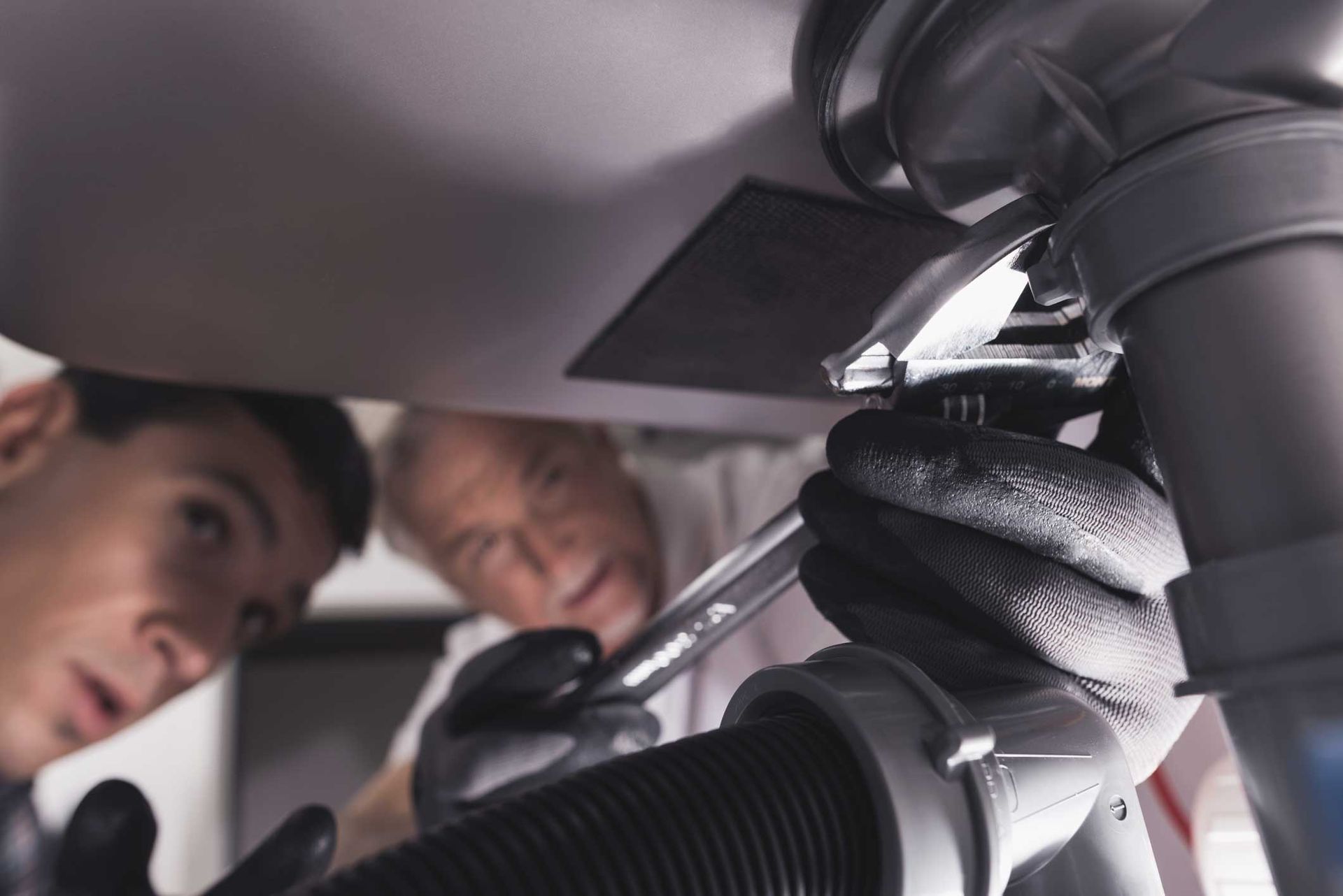 Two plumbers looking under a sink while one uses a spanner on a pipe.