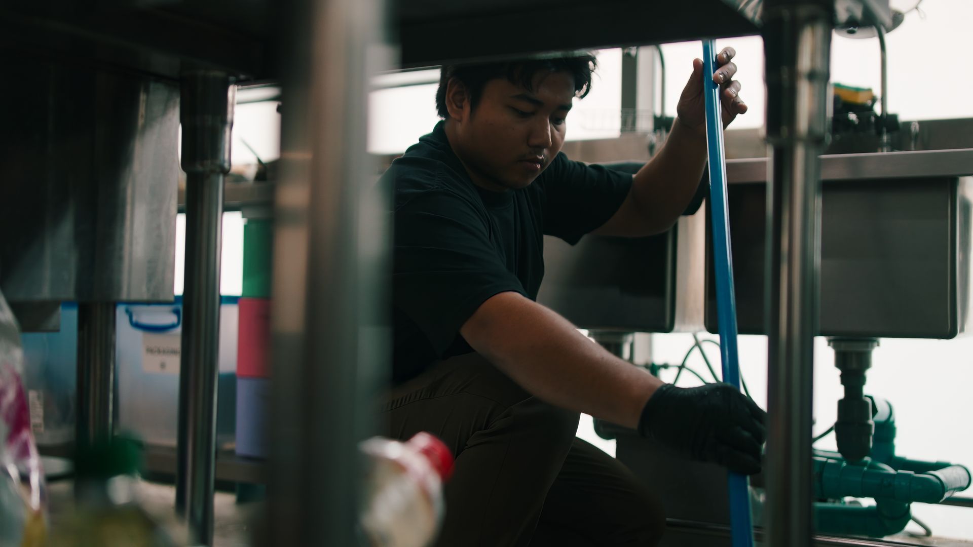 A plumber is performing commercial plumbing services, working under a stainless steel sink.