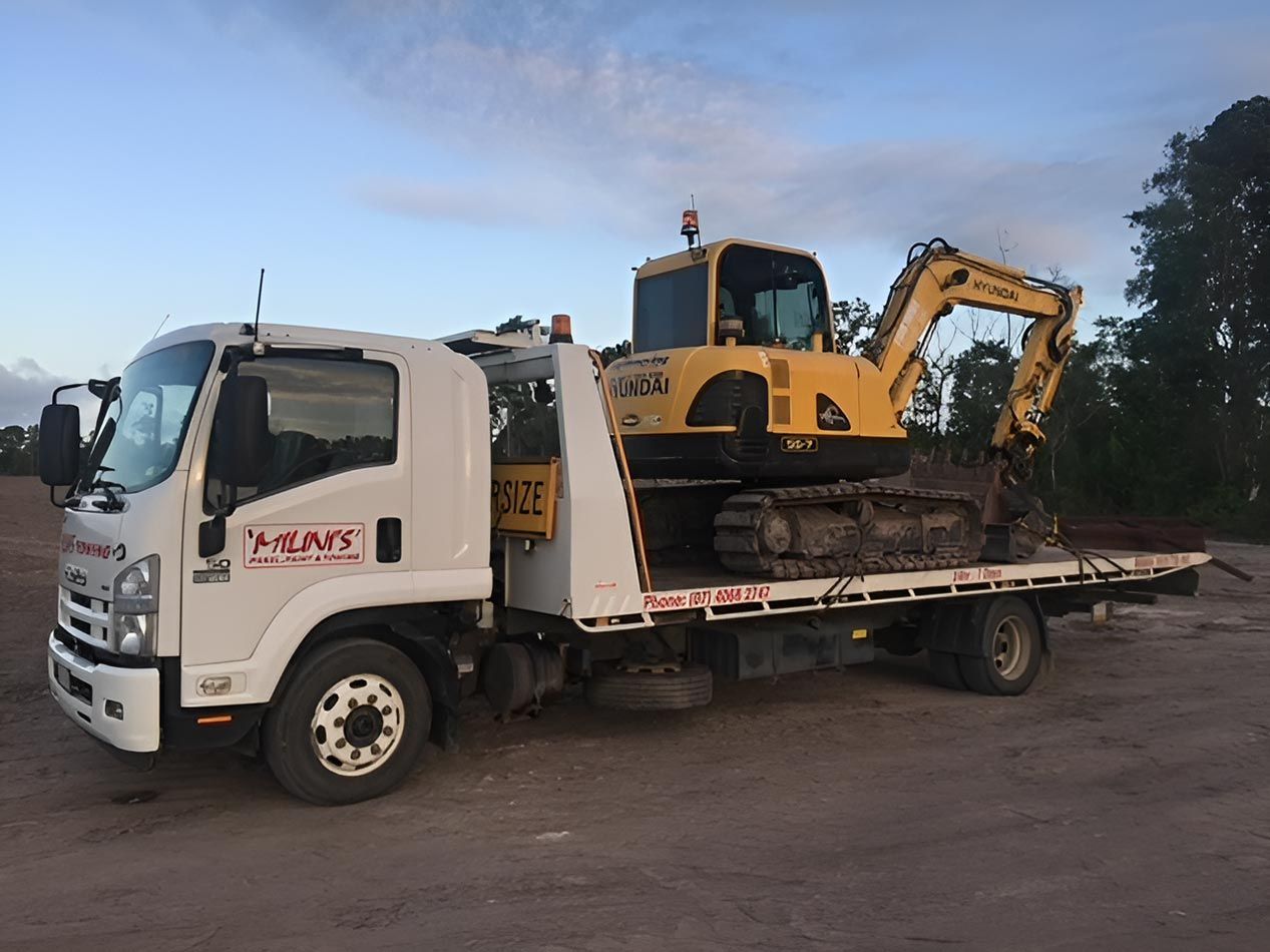 A Tow Truck Is Carrying A Yellow Excavator On The Back Of It — Milini's Panel, Paint & Towing in Mission Beach, QLD