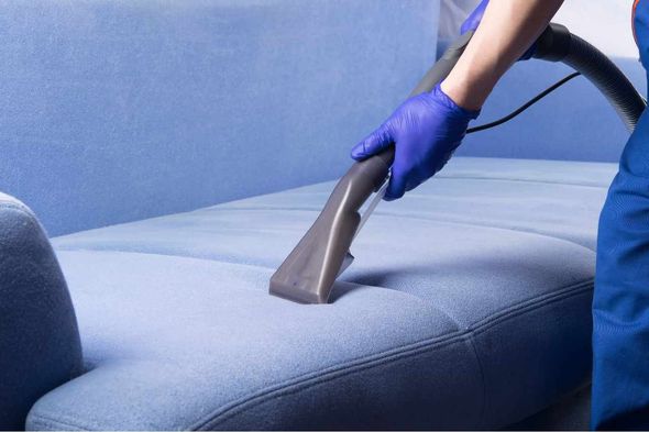 A person is cleaning a blue couch with a vacuum cleaner.