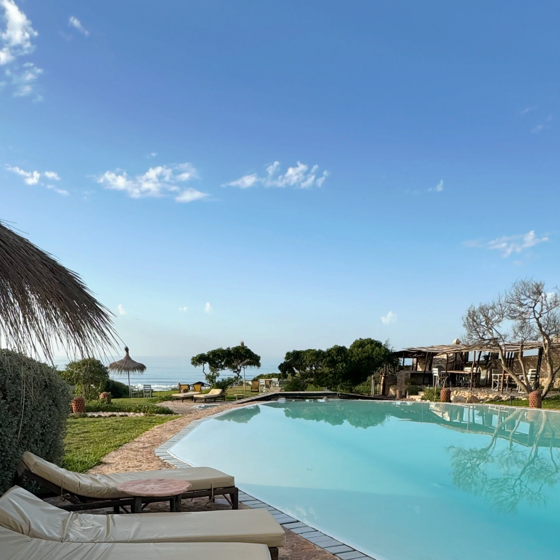 Poolside view with clear blue water, ocean in the distance, blue sky, lounge chairs.