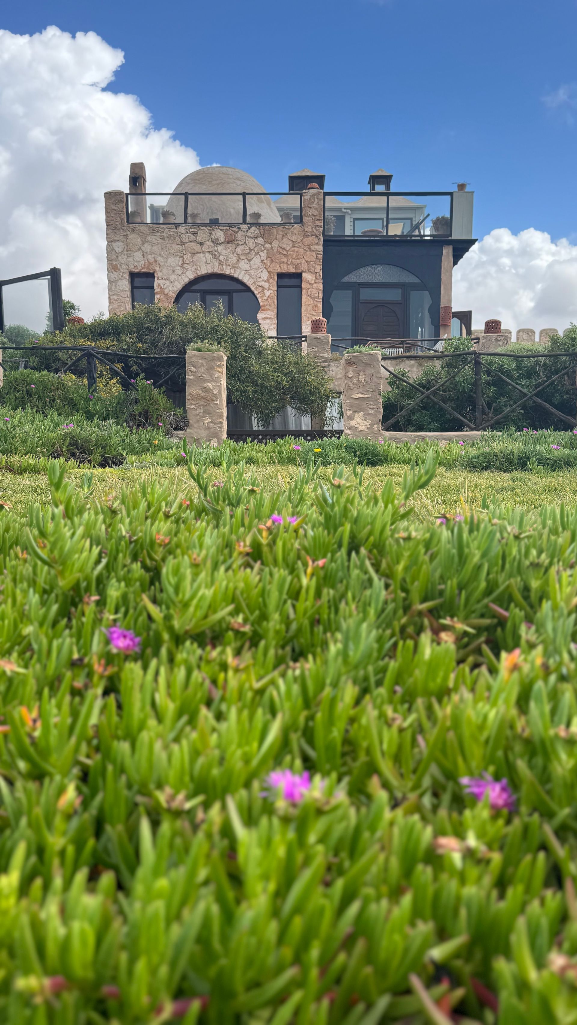 Stone building with dome and modern glass extension, surrounded by green plants and a blue sky.