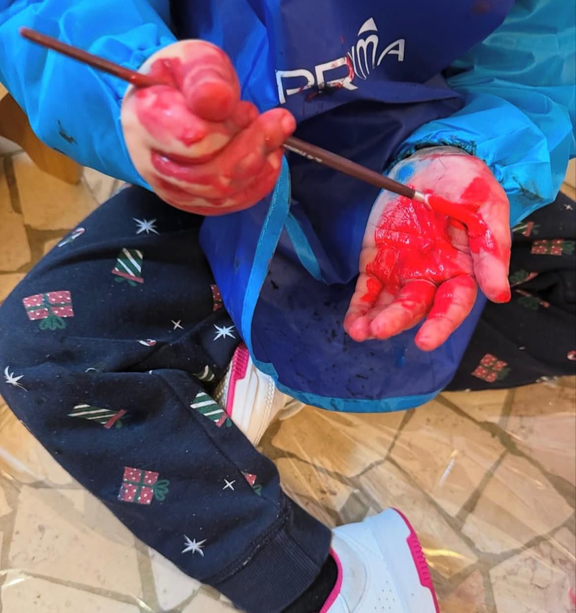 Bambino ricoperto di vernice rossa, con in mano un pennello, che indossa un camice blu e pantaloni con stampe natalizie.