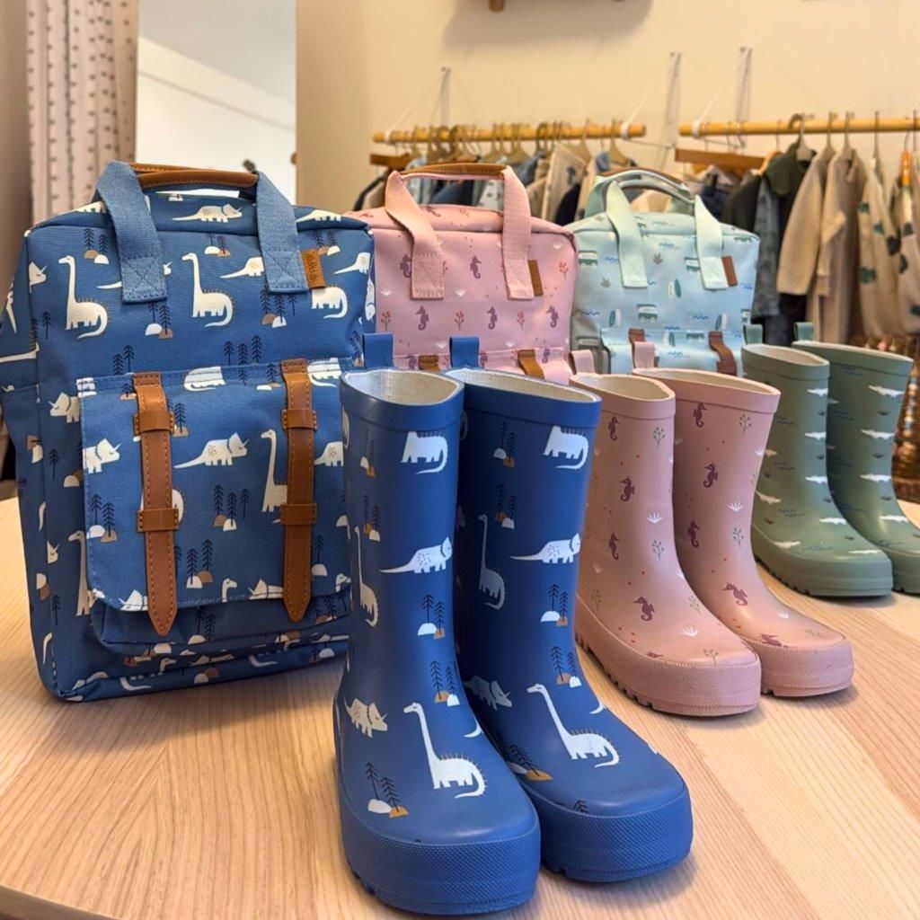 Zaini e stivali da pioggia per bambini a tema dinosauri, nei colori blu, rosa e verde, esposti su un tavolo.