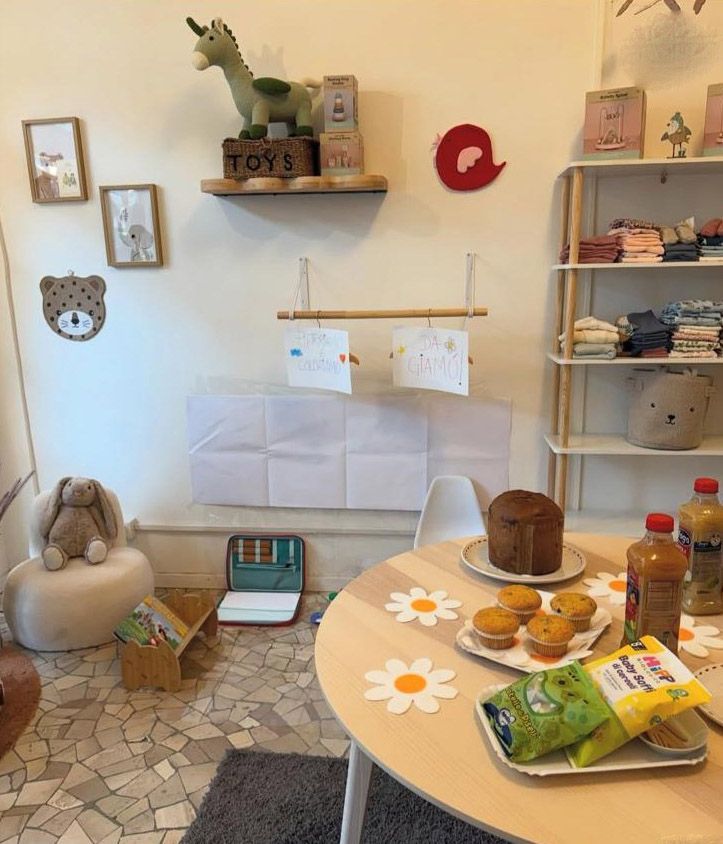 Interno di un negozio per bambini: giocattoli, tavolo con snack, scaffali con vestiti e opere d'arte alle pareti.