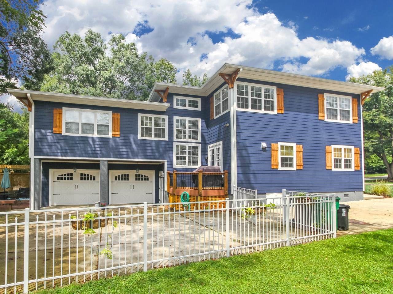 Blue two-story house with white garage doors, wooden shutters, and a white picket fence on a sunny day.