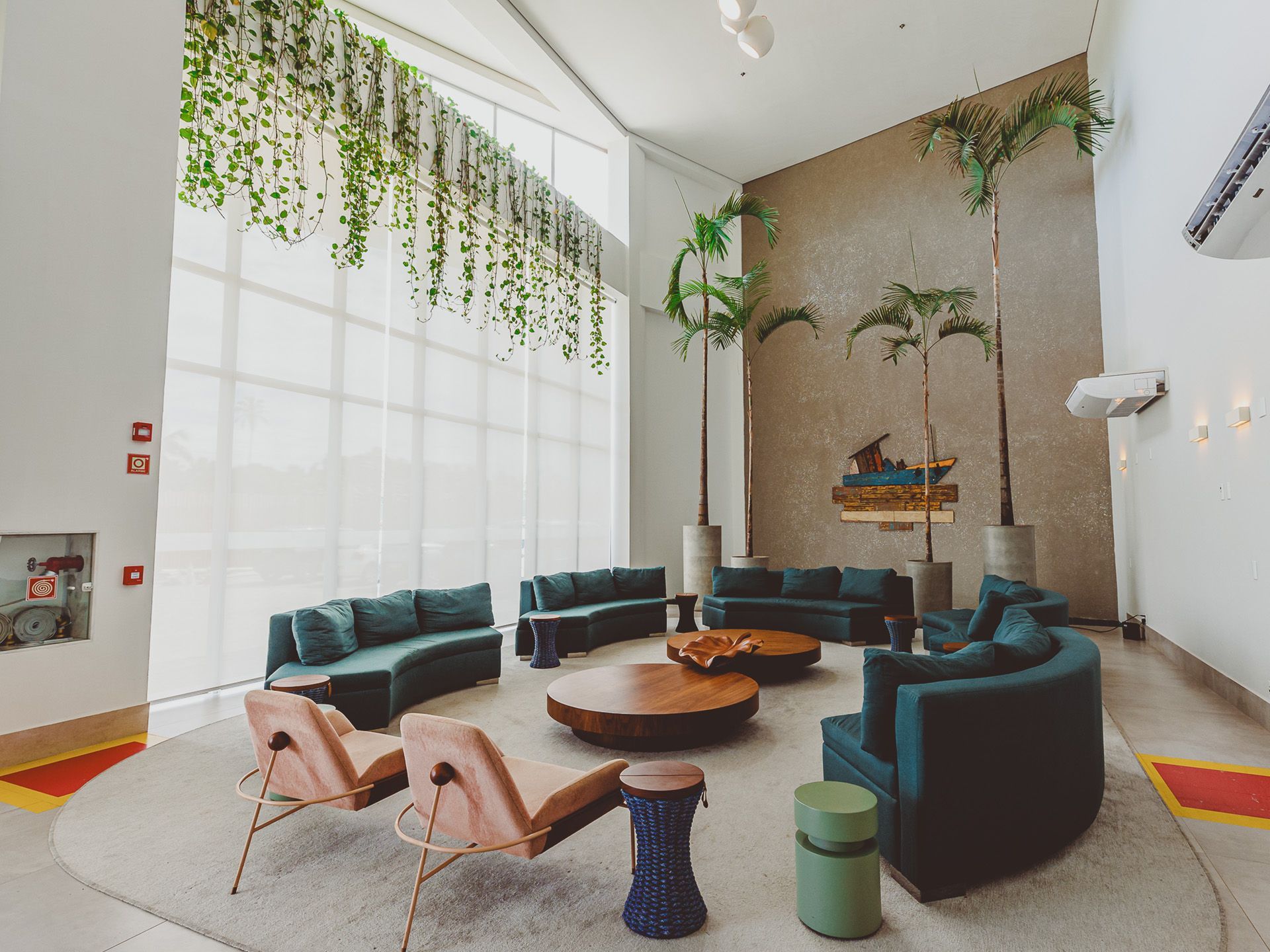 Sala de estar com sofás azul-petróleo, mesa de centro redonda de madeira e plantas penduradas em uma parede branca.