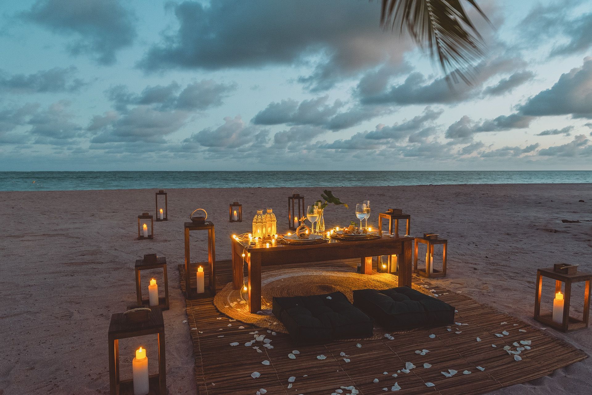 Jantar romântico na praia ao anoitecer com lanternas, mesa, travesseiros e vista para o mar.