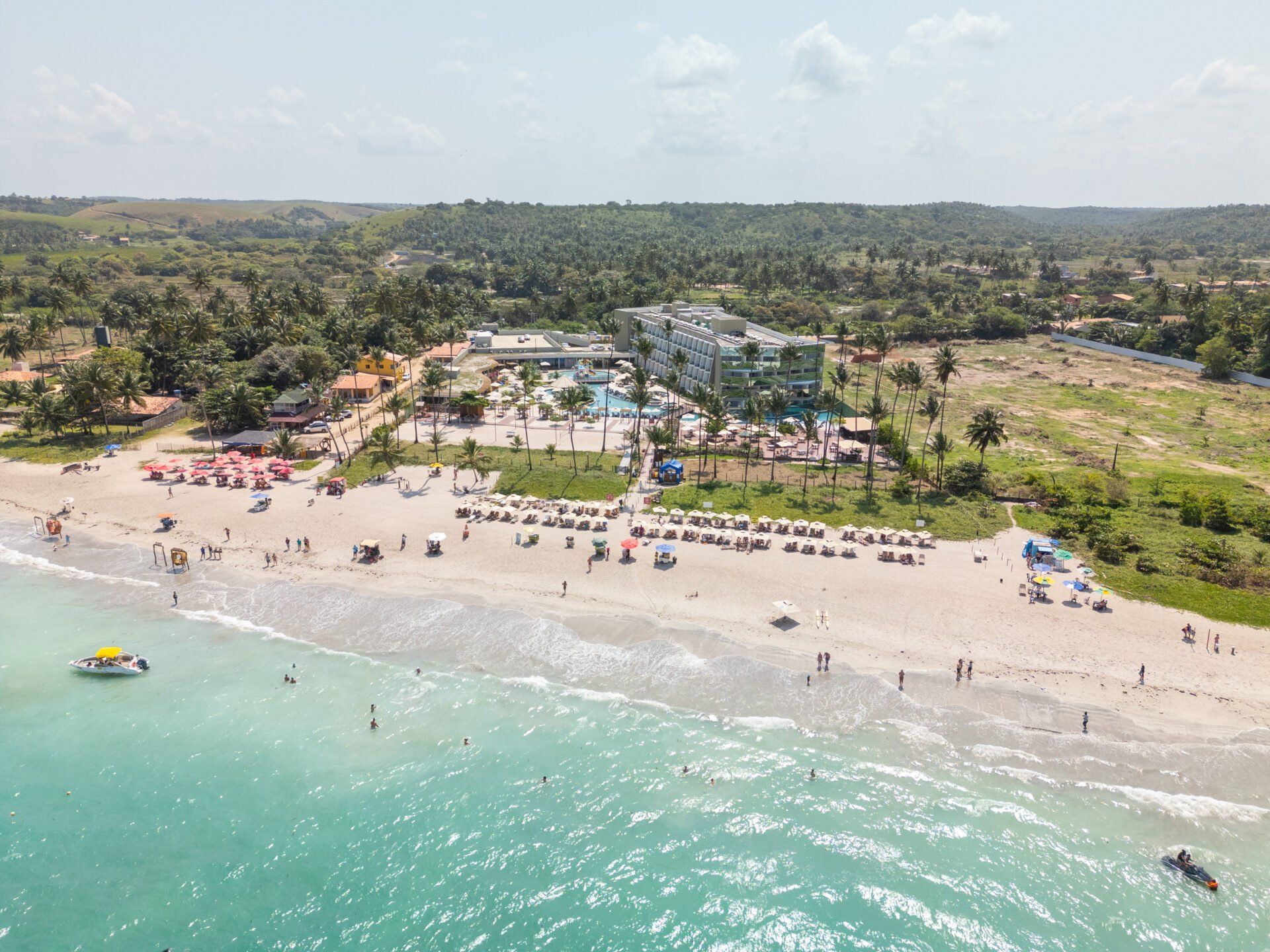 Praia com águas azul-turquesa, areia fina, hotel e pessoas. Palmeiras e céu azul visíveis.