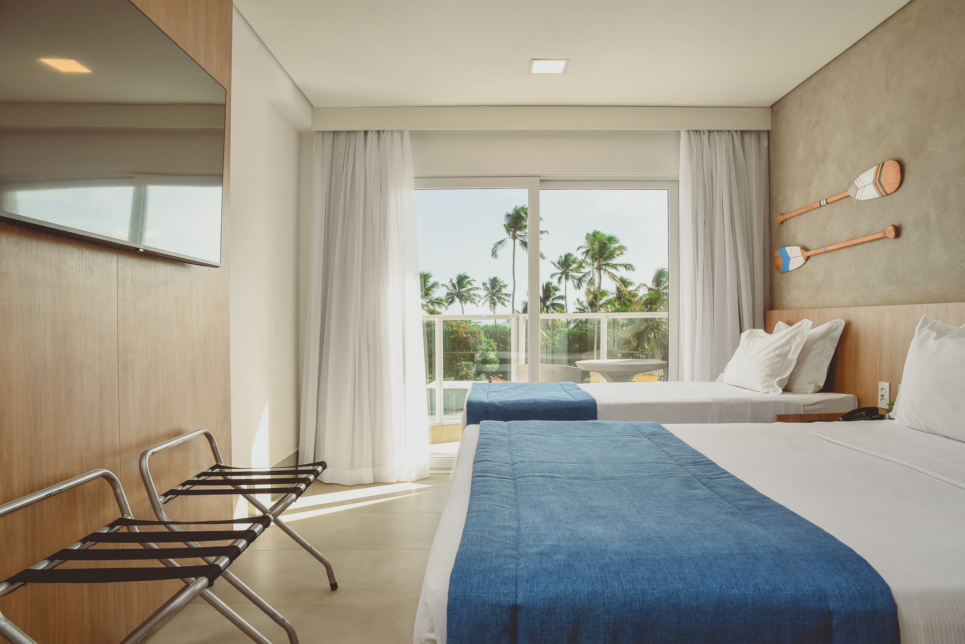 Quarto com cama, vista para o mar e varanda. Paredes brancas, detalhes em azul e palmeiras.