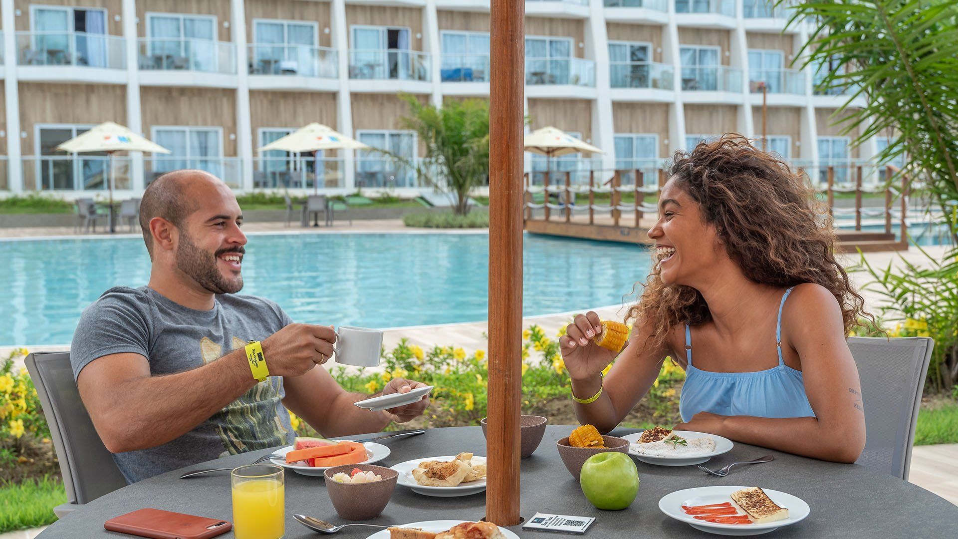 Um casal ri durante o café da manhã em uma mesa perto de uma piscina, com um resort ao fundo.