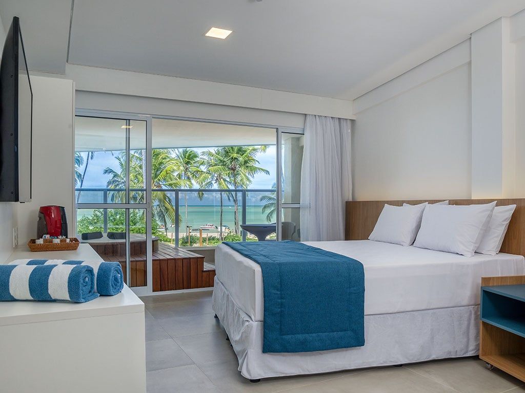 Quarto com cama, vista para o mar e varanda. Paredes brancas, detalhes em azul e palmeiras.