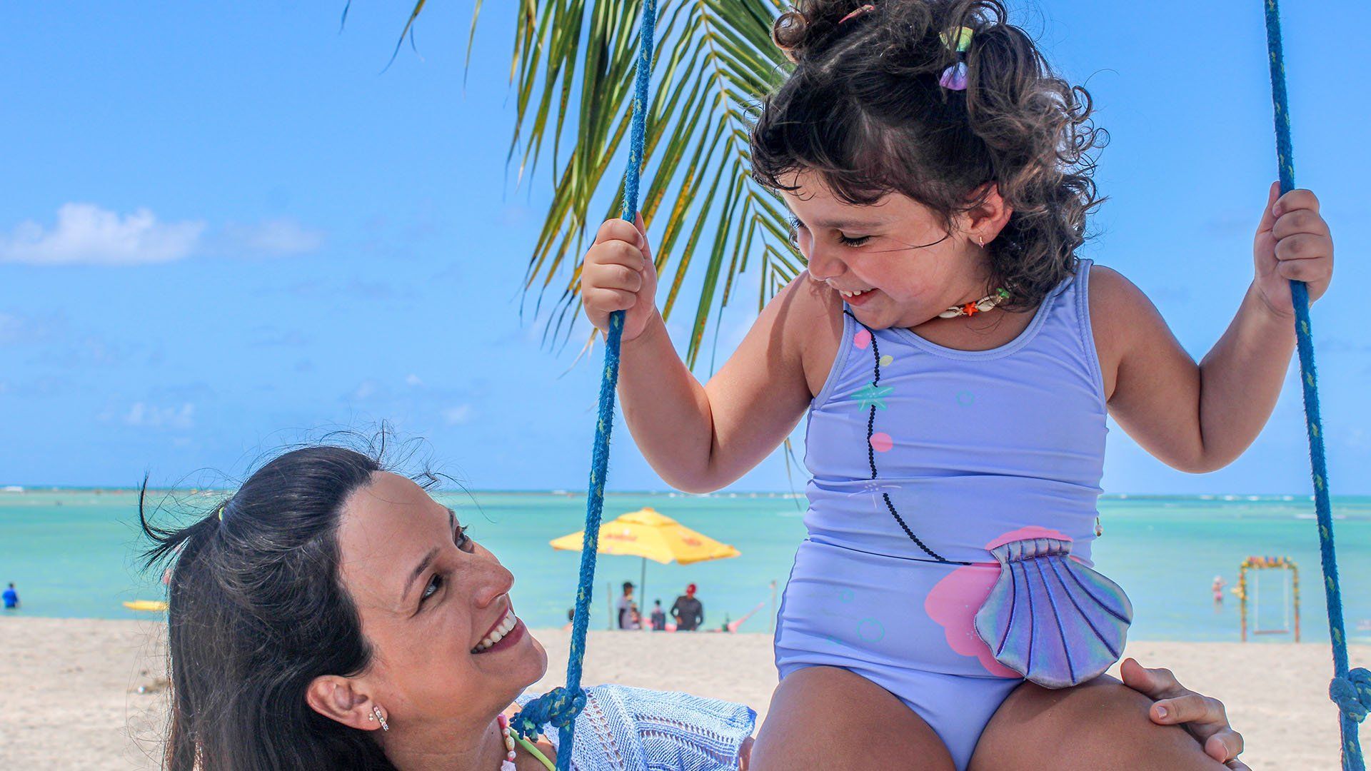Mulher sorrindo para uma criança em um balanço de praia; dia ensolarado, criança em traje de banho, céu azul.