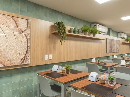 Interior do restaurante: mesas de madeira, parede de azulejos verdes, painéis de madeira com plantas e obras de arte, cadeiras cinza.