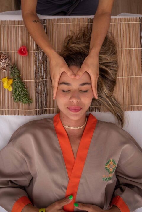 Mulher recebendo massagem na cabeça, olhos fechados, usando roupão de spa. Mãos na testa.