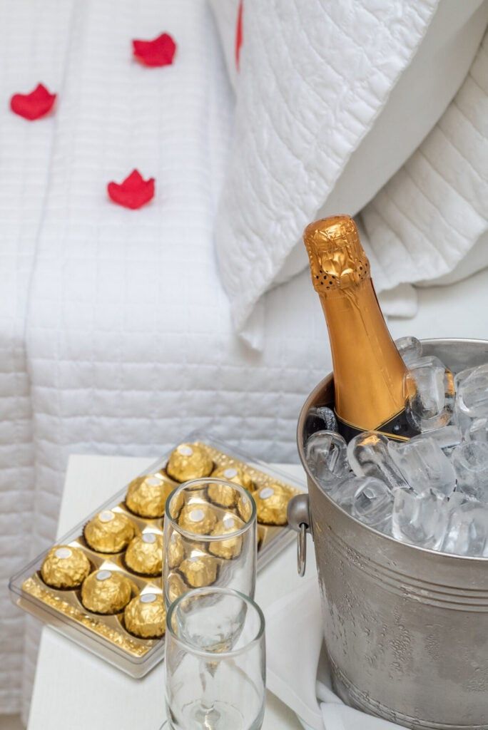 Champanhe em balde de gelo com chocolates e taças sobre uma mesa branca ao lado de uma cama com pétalas de rosa.