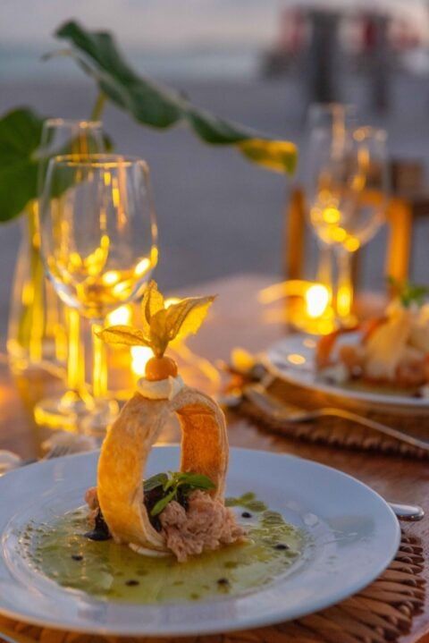 Jantar romântico na praia: comida gourmet, taças de vinho, velas, pôr do sol ao fundo.