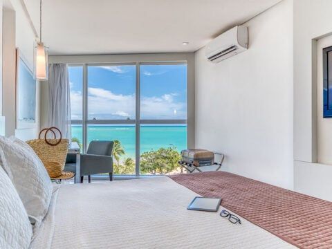 Quarto iluminado com uma janela grande com vista para o oceano azul-turquesa. Paredes brancas, cama com cobertor rosa.