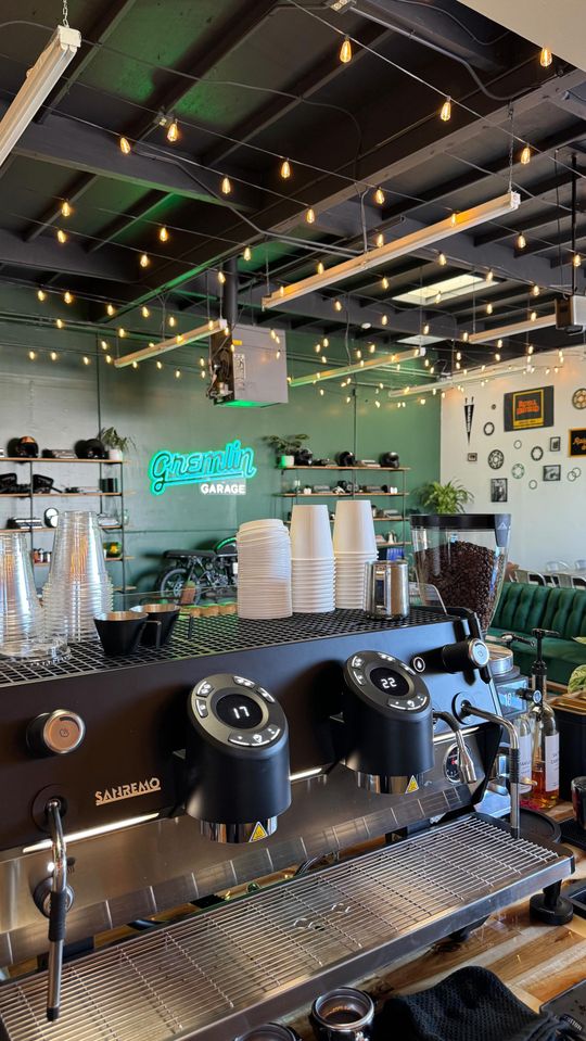 A close-up of a professional espresso machine in a modern, green-walled coffee shop with string lights on the ceiling.