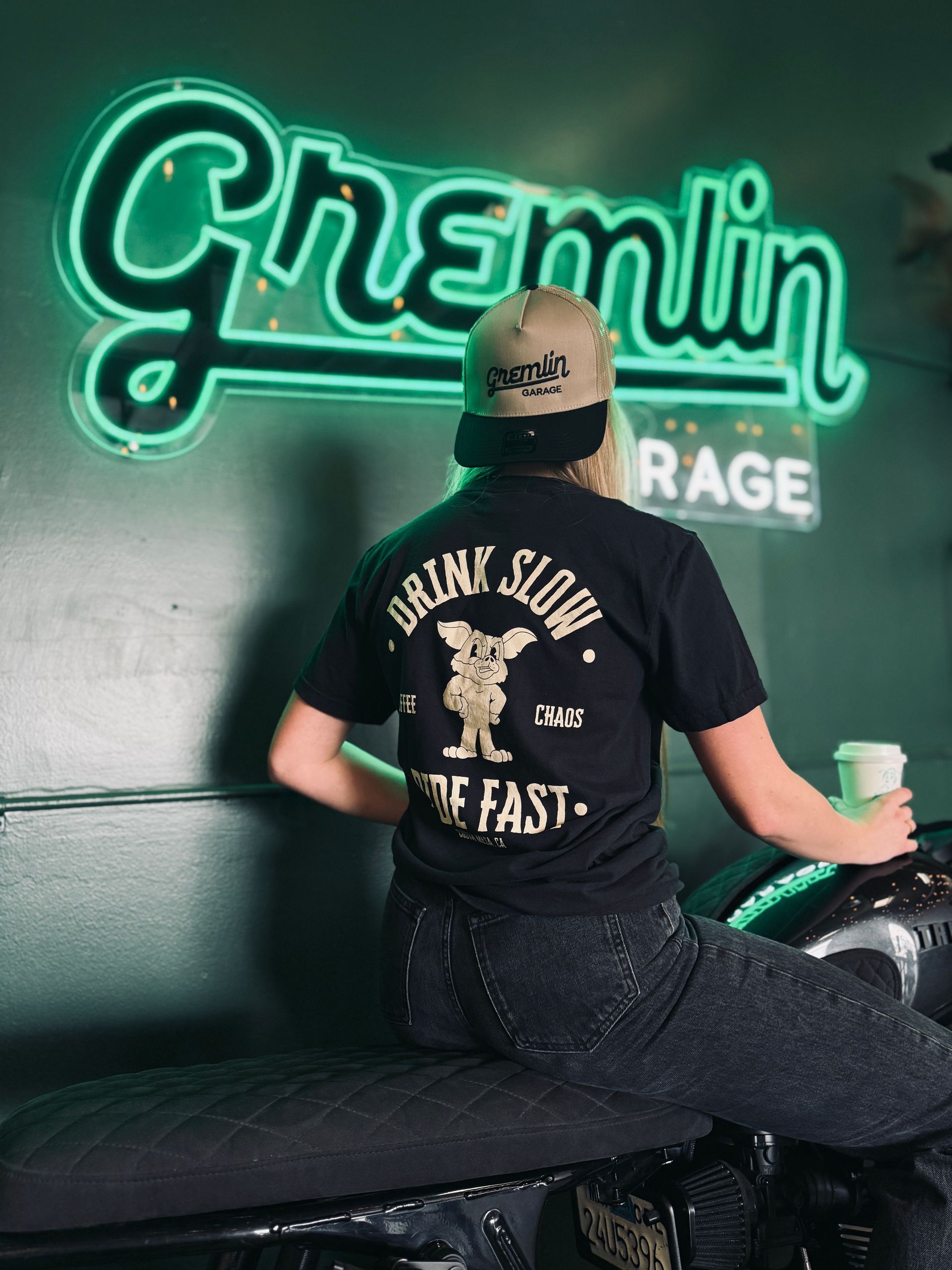 A woman is sitting on a motorcycle in front of a neon sign that says chemin garage