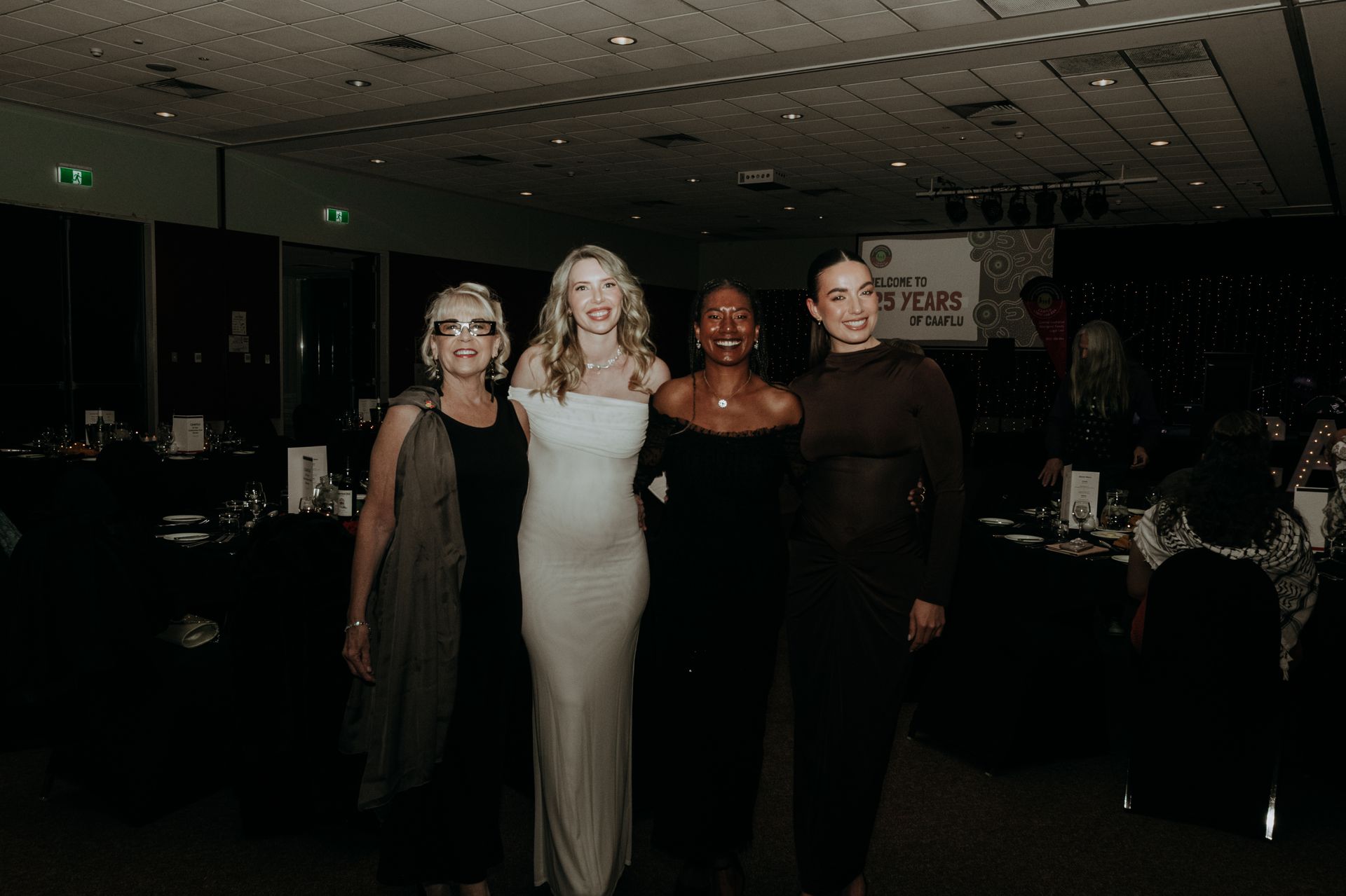Four women pose at an event. One wears white, others in black dresses. Dimly lit venue.