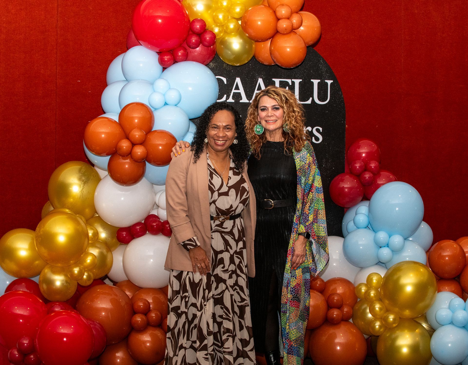 Two women smiling, posing by colorful balloons at an event.