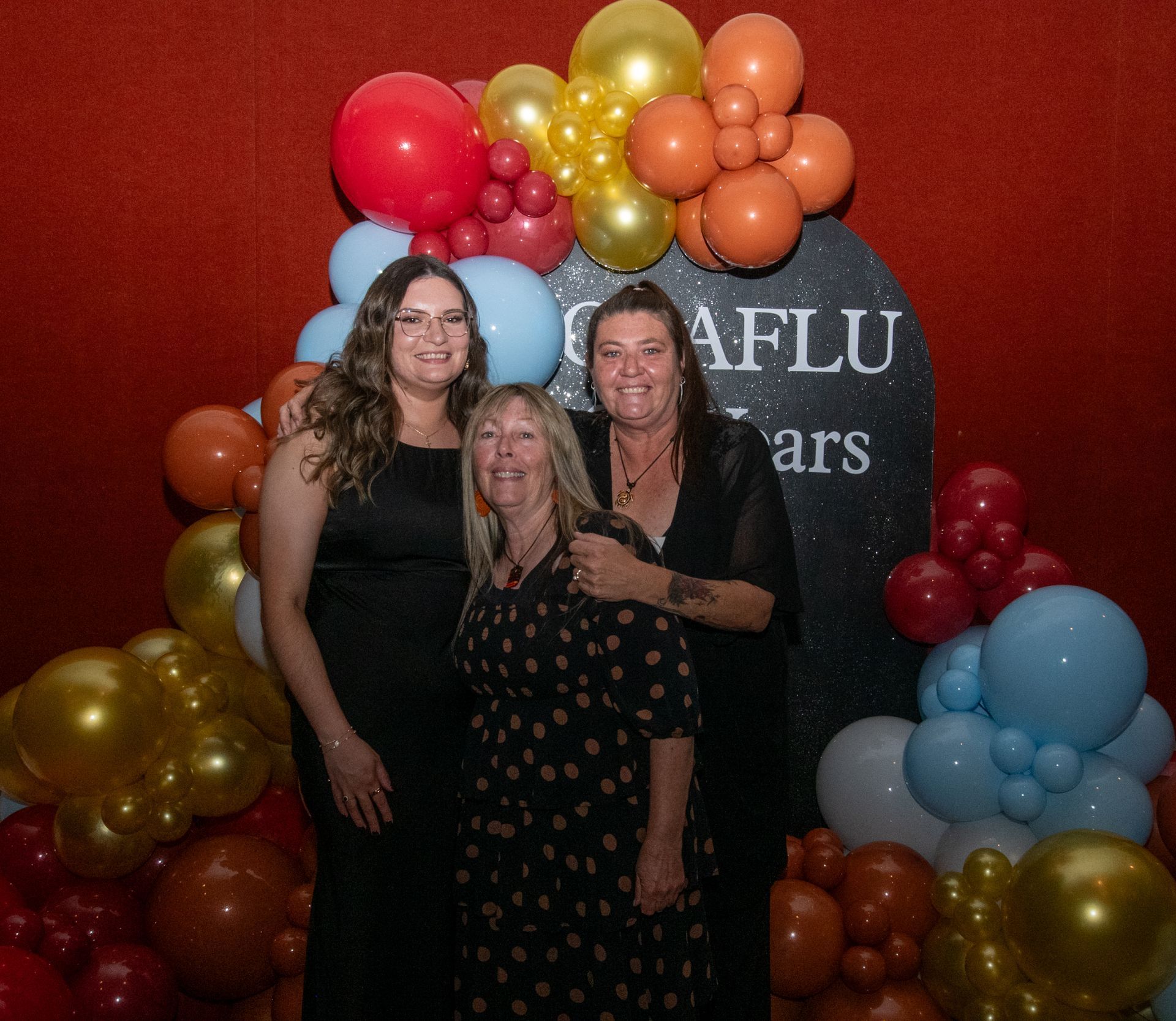 Three women posing, smiling in front of a balloon arch at an event.