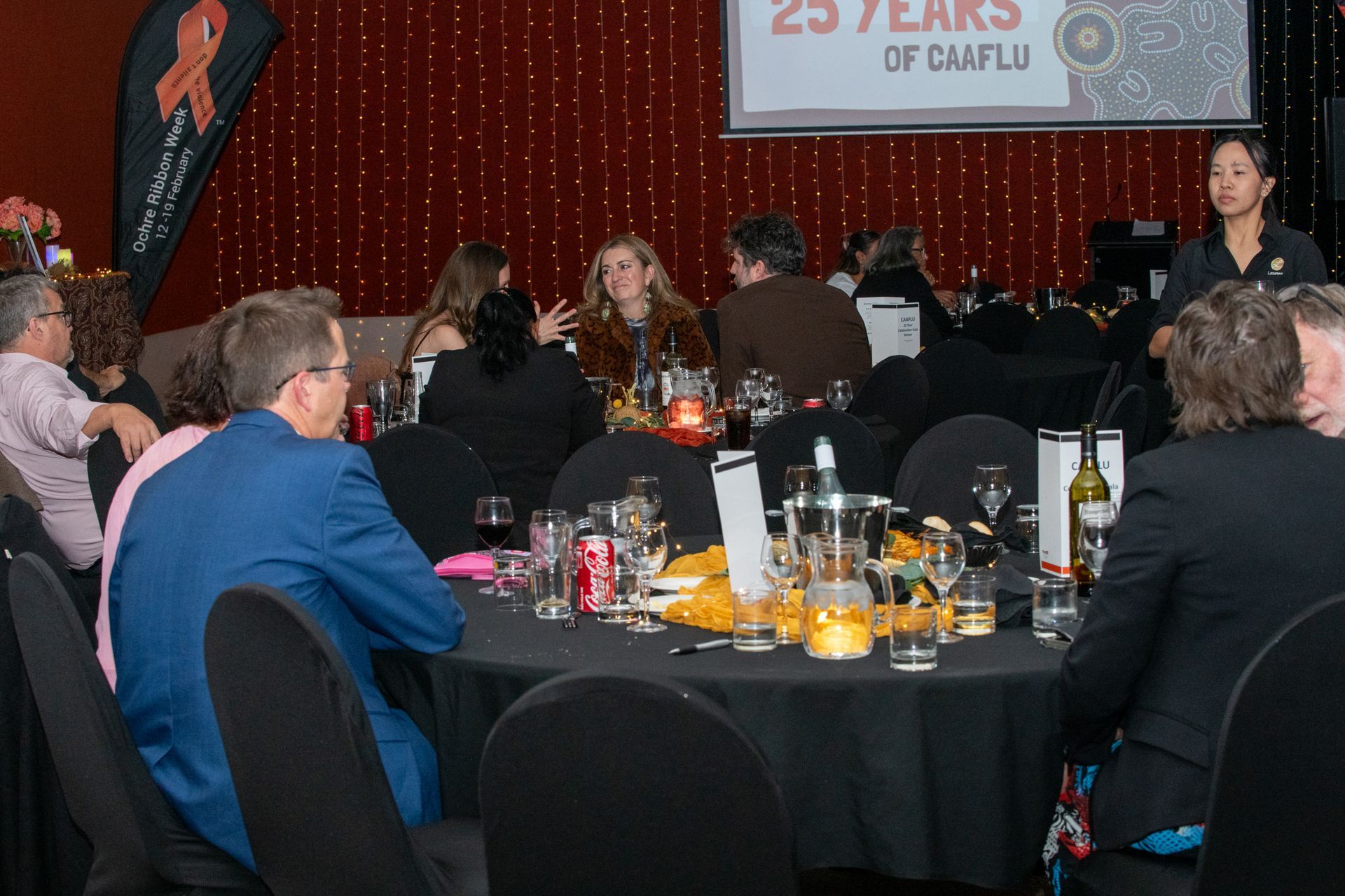 People at a formal event seated at round tables, with a server, a backdrop with a banner and twinkling lights.