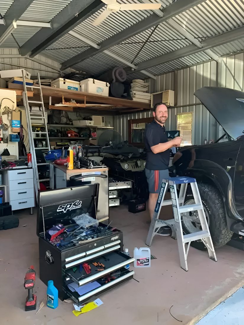 A Man Is Standing On A Ladder In A Garage Working On A Car — Deri's Mobile Mechanical In Parap, NT