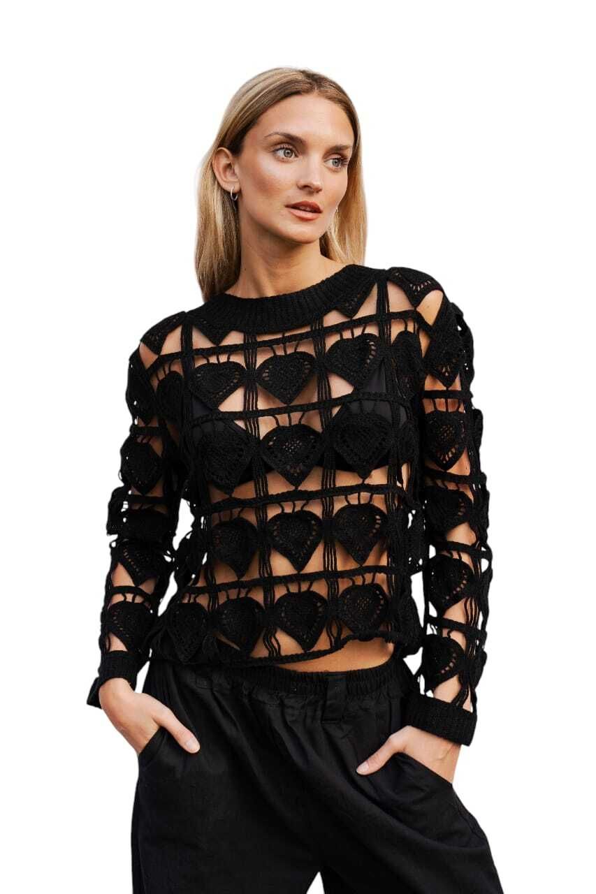 Mujer con suéter y pantalones negros con estampado de corazones, manos en los bolsillos, mirando fuera de cámara.