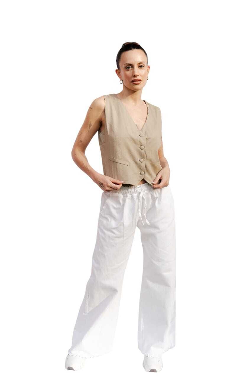 Mujer con top color canela sin mangas y pantalones blancos con cordón, de pie, mirando hacia adelante.