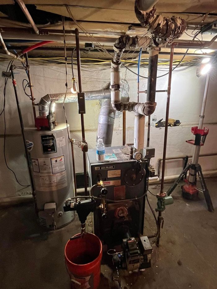 A water heater is sitting in a basement next to a red bucket.